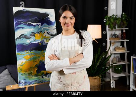 A young woman stands confidently in her bright home studio filled with colorful paintings. Stock Photo