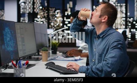 Tech startup worker drinking coffee, developing AI algorithms in high rise office. Caffeinated programmer energetically debugging neural network brain models on screens Stock Photo