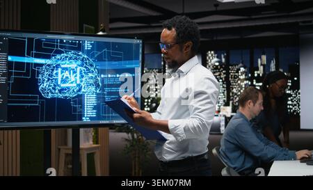 Manager in office writing on notepad, taking notes of artificial intelligence system bugs to fix. Supervisor looking at large screen, documenting neural network errors during troubleshooting Stock Photo