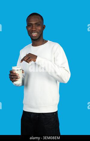 Young man smiling, holding a reusable coffee cup, pointing at it on a blue background Stock Photo