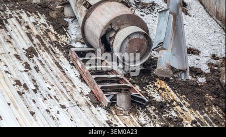 Workers are handling construction materials and equipment on the ground at a site. A ladder rests against a structure. Dust and dirt cover the area. A large container lies on its side. Stock Photo
