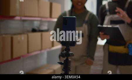 Warehouse logistics coordinator and colleague using smartphone placed on tripod to make training video for interns. Employees film themselves in fulfillment center showing trainees how to seal boxes Stock Photo