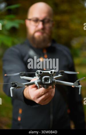 Man holds drone toward camera with focus on device. Quadcopter in sharp focus with blurred bearded man in background, shallow depth of field in park, concept of modern technology and hobby Stock Photo