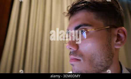 Male face of manager in glasses with reflection of screen lights working on laptop in home interior. Focused young businessman using computer in Stock Photo