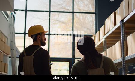 Meticulous trainee walking through warehouse rows scanning cardboard boxes on shelves under team leader guidance, using digital devices to update info before sending parcels for delivery Stock Photo