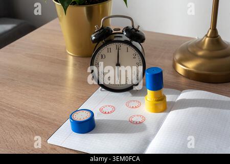 Black vintage alarm clock on wooden desk next to colorful clock stamps, open notebook with red clock face prints and golden lamp. Concept of time Stock Photo