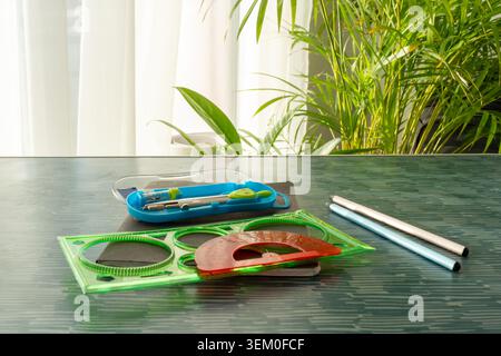 Green geometric ruler, red protractor and compass set in blue case on dark desk with pencils near window with tropical plants. Concept of geometry Stock Photo