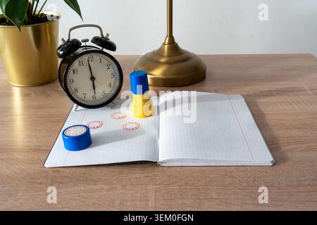 Clock face rubber stamps with red ink prints on open notebook next to black alarm clock, golden lamp and green plant on wooden desk. Time learning Stock Photo