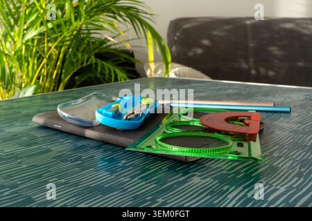 Colorful geometry set with green ruler, red protractor and compass in blue case on dark table with tropical plant background. Concept of school Stock Photo