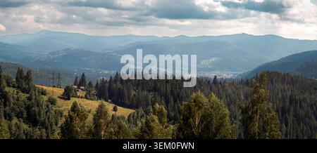 Wide panoramic view of a mountain valley with dense evergreen forests, rolling hills and distant village under a dramatic cloudy sky. Natural summer l Stock Photo