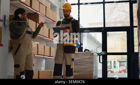 Chief supply chain officer and employee preparing warehouse orders for delivery, receiving phone call in depot while scanning labels on cardboard boxes parcels to be shipped Stock Photo