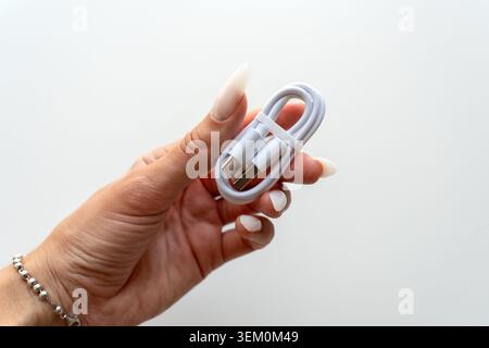 Horizontal photo of a female hand holding a neatly coiled white USB Type-C cable secured with a rubber band on a white background Stock Photo