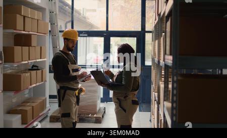 Warehouse chief supply chain officers inputing updated products info on online shopping website, walking through storeroom aisles inspecting labels on wares, crosschecking with inventory list Stock Photo