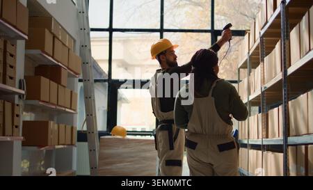 Meticulous warehouse inventory counter scanning cardboard boxes merchandise on shelves under team leader guidance, using digital devices to update prices for internet ecommerce website, hip level shot Stock Photo