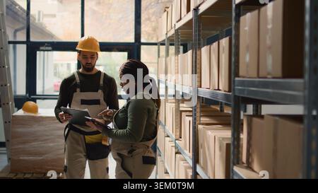Dolly out shot of warehouse staff inputing updated products info on online shopping website, walking through distribution center aisles inspecting labels on wares, crosschecking with inventory list Stock Photo