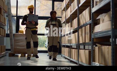 Experienced employees walking through warehouse aisles checking labels on cardboard boxes sitting on shelves, inputing updated merchandise info on online ecommerce website Stock Photo
