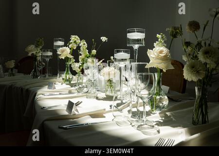 Luxury indoor wedding reception table set with all-white florals, floating candles, crystal wine glasses and place cards in dramatic moody light Stock Photo