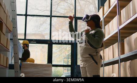 African american employee in warehouse wearing VR headset, using technology to visualize how to best stack cardboard boxes on shelves to make room for more. Worker in storage room uses virtual reality Stock Photo