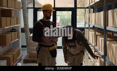 Employee and helpful colleague inputing updated products info on online shopping website, walking through warehouse storage center rows checking labels on cardboard boxes sitting on shelves Stock Photo