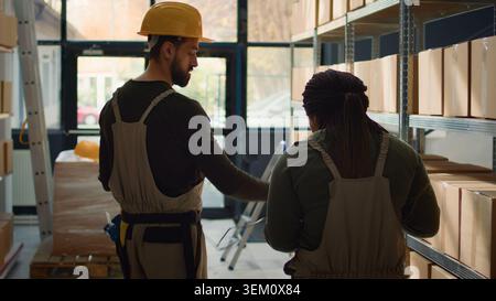 Meticulous employee walking through warehouse aisles scanning cardboard boxes on shelves under team leader guidance, using digital devices to update info before sending packages for shipping Stock Photo