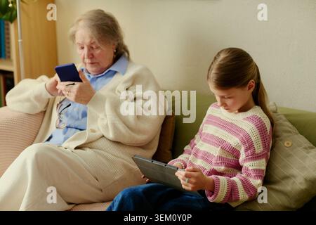 Senior woman sitting with granddaughter, using smartphone while girl using tablet at home. Illustrating family caregiving, digital learning, elder support, leisure, medium shot Stock Photo