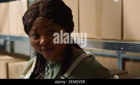 Portrait of smiling warehouse head of logistics holding digital tablet, checking packaging, ensuring flawless operations while employee in background handles packaged merchandise in cardboard boxes Stock Photo