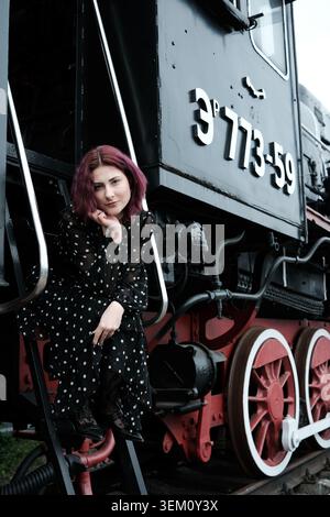 A woman with pink hair sits on steps of a vintage steam train, radiating charm and style. Stock Photo