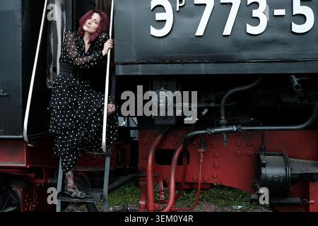 Woman in a polka dot dress elegantly poses next to a classic train, radiating charm. Stock Photo