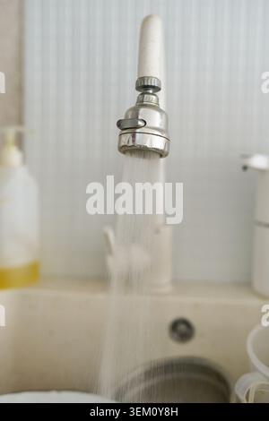 Kitchen Faucet Spraying Steady Stream Of Water Into Sink Basin Filled With Dishes And Containers On Countertop In Domestic Interior With Tiled Backsplash And Soap Dispenser Visible. Stock Photo