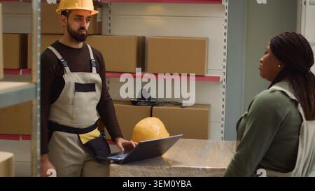 Tracking shot of worker and colleague inputing updated products info on online shopping website, walking through warehouse depository rows checking labels on goods sitting on shelves Stock Photo