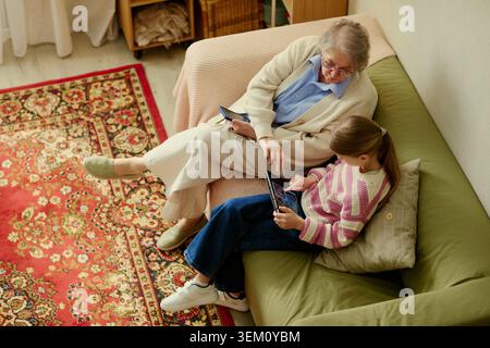 Senior woman and granddaughter using tablet together at home, sharing digital caregiving support and family connection. Useful for elder care, intergenerational learning, home assistance marketing Stock Photo