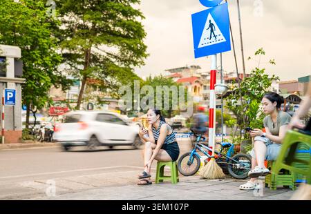 Vietnam-June 11 2025:Near to the Old Quarter and beside the Red River,a woman working in a street cafe uses her phone by the roadside,next to city tra Stock Photo