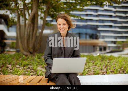 A woman sits on a bench in an outdoor area. She uses a laptop while smiling. Behind her is a modern building and trees. It is a sunny day with clear s Stock Photo