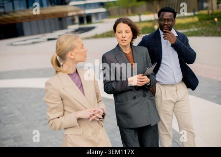 Three individuals stand outside a building. Two women and one man are engaged in a conversation. One woman holds a tablet while the others listen and Stock Photo