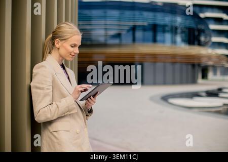 A businesswoman stands against a pillar while using a tablet. She appears focused on her device. The setting shows a modern building in a city during Stock Photo