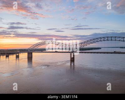 A steel arch bridge spans the wide Mississippi River at sunset, with vehicles crossing and colorful clouds reflecting on the water. Stock Photo