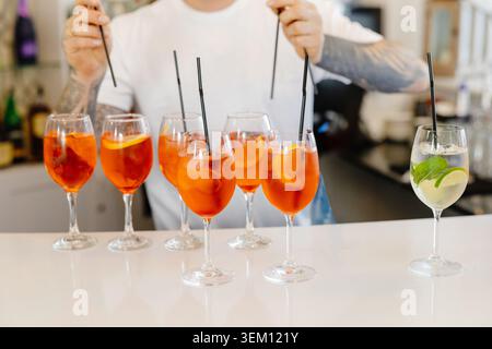 Bartender adding black straws to glasses of aperol spritz and a hugo cocktail on a bar counter Stock Photo