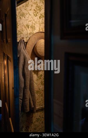 Straw hat and grey cardigan hanging on hooks beside floral wallpaper, glimpsed through a doorway in warm atmospheric light Stock Photo