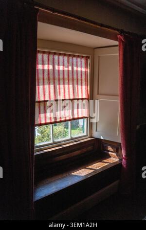 Sunlight filtering through striped red roller blind at a rustic window with dark wood seat and draped curtains Stock Photo