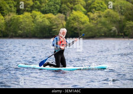 Elderly man with white beard paddleboarding on a lake, wearing wetsuit and life jacket, paddling a Rockit inflatable SUP board Stock Photo