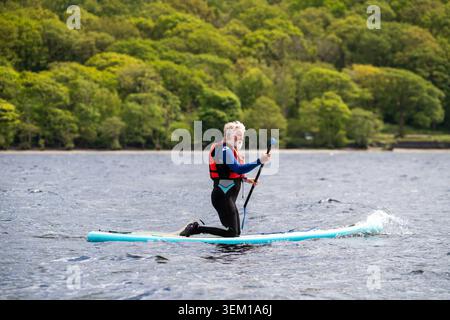 Older man with white beard paddleboarding on a calm lake wearing wetsuit and red life jacket, lush green trees behind Stock Photo