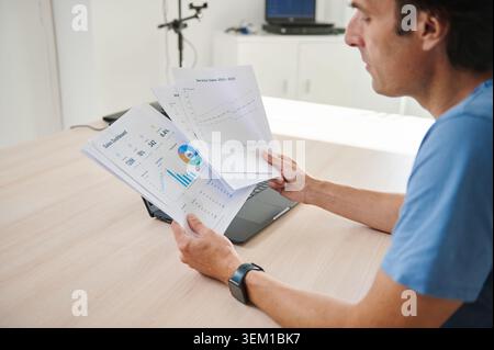 Man holding printed sales reports and charts while working on a laptop, analyzing data and trends in a bright office environment conveying focus, deci Stock Photo