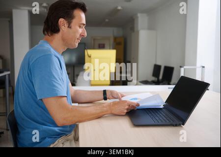 Man seated at office desk, reviewing printed reports and charts while using a laptop - calm focused small-business or remote-work scene with modern wo Stock Photo