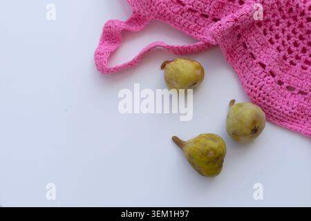 Bright top view of three ripe green figs arranged near a pink crochet string bag on a white background with large copy space. Stock Photo
