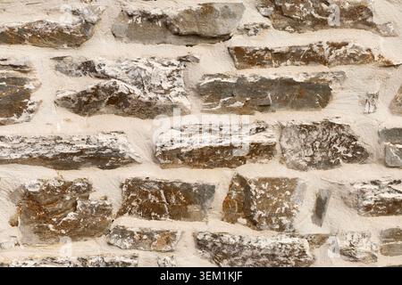 Flat lay view of an old rustic wall made from coarse brown rocks and grey stone blocks embedded in textured white plaster mortar. Stock Photo