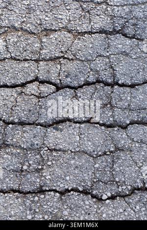 Vertical top view of a heavily cracked, weathered grey asphalt pavement texture showing detailed fractures and embedded stones. Stock Photo