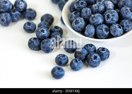 High angle view of ripe juicy blueberries scattered on a light beige studio background next to a full ceramic bowl with selective focus. Stock Photo