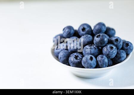 A side view close-up shot of a white ceramic bowl filled to the brim with ripe juicy blueberries, placed on a neutral light background with a large em Stock Photo
