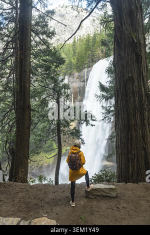 Solo hiker wearing a yellow rain jacket and backpack stands on a viewing point, gazing at the powerful flow of vernal fall surrounded by lush forest a Stock Photo