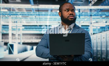 Happy programmer in server hub using laptop to do maintenance tasks on infrastructure machines. Cheerful man in modern high tech data center configuring server settings with notebook, camera B Stock Photo